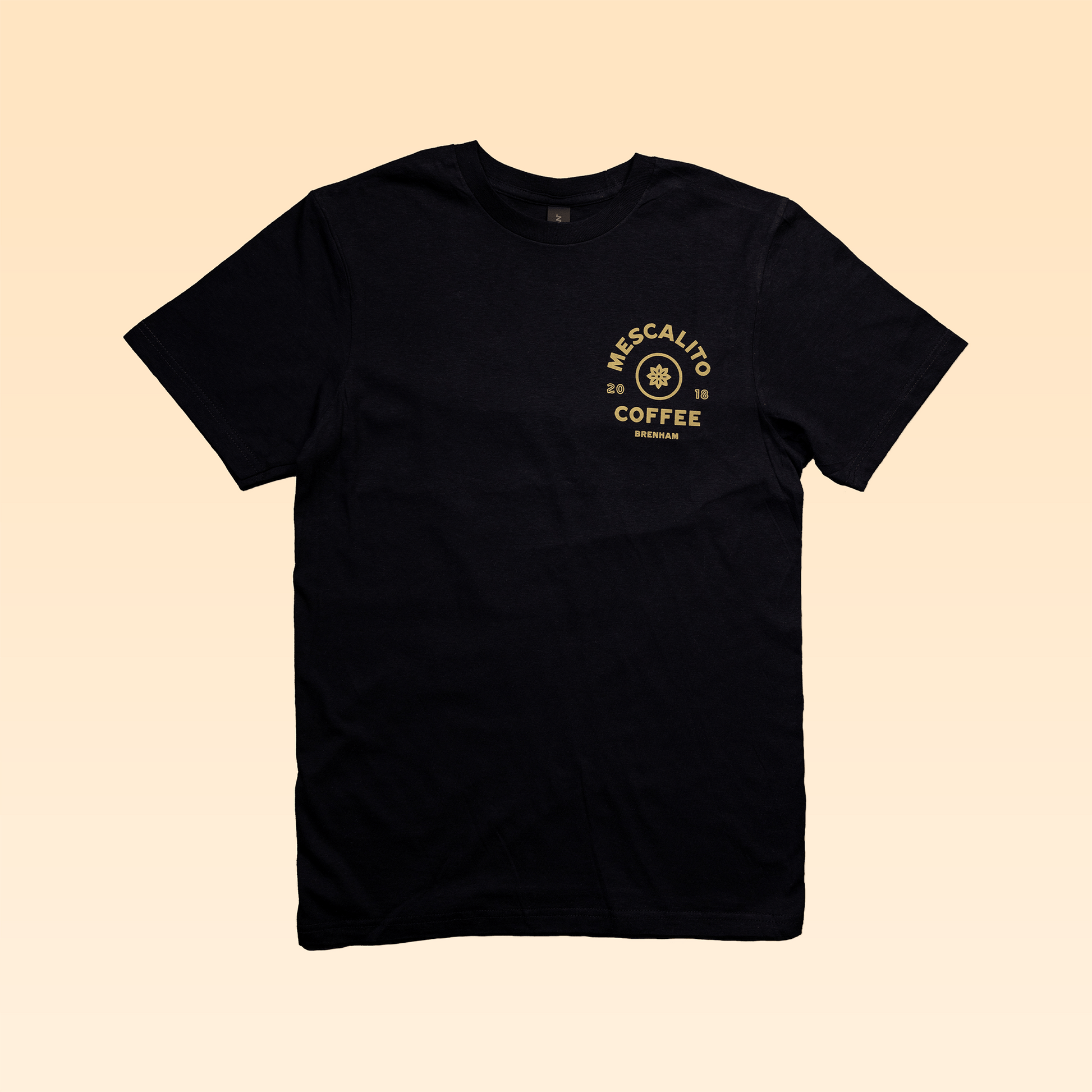The Mescalito Coffee Shop Shirt by Mescalito Coffee features the signature yellow logo on the upper left chest, laid flat on a light beige background—inspired by Brenham’s favorite spot.