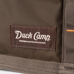 Close-up of Duck Camp's Mesh Gear Tote, a brown outdoor bag featuring a stitched "Duck Camp Premium Outdoor Goods" label.