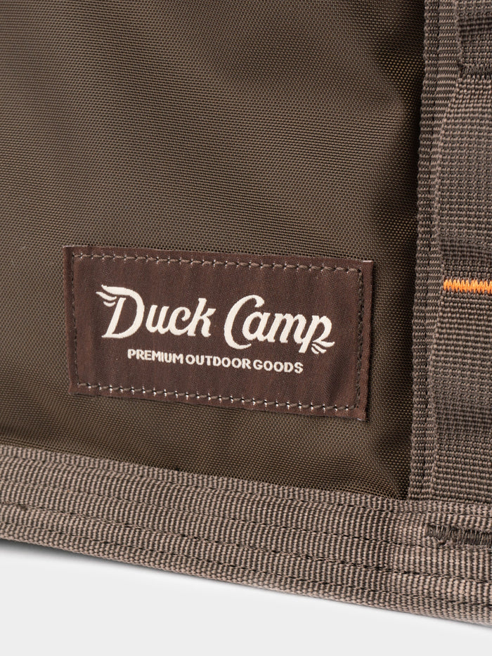Close-up of a brown fabric label with "Duck Camp Premium Outdoor Goods" stitched onto the water-resistant Mesh Gear Tote by Duck Camp, perfect for organizing your outdoor gear.