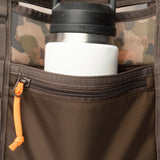 A Duck Camp Mesh Gear Tote in brown camouflage holds a stainless steel Yeti water bottle with a black lid, placed in its front zippered pocket.