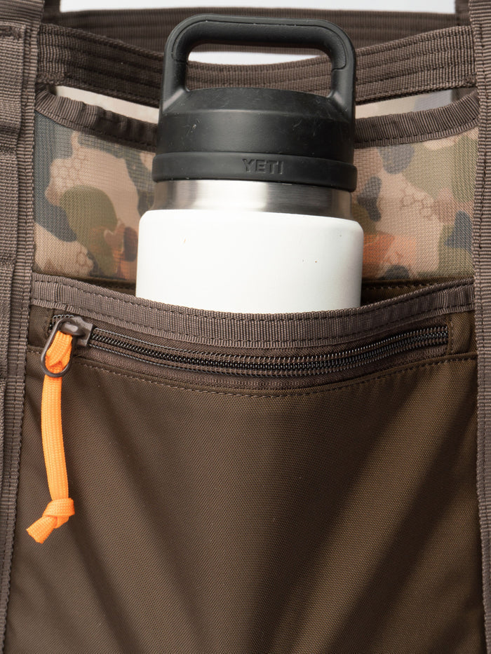 A Duck Camp Mesh Gear Tote in brown camouflage holds a stainless steel Yeti water bottle with a black lid, placed in its front zippered pocket.