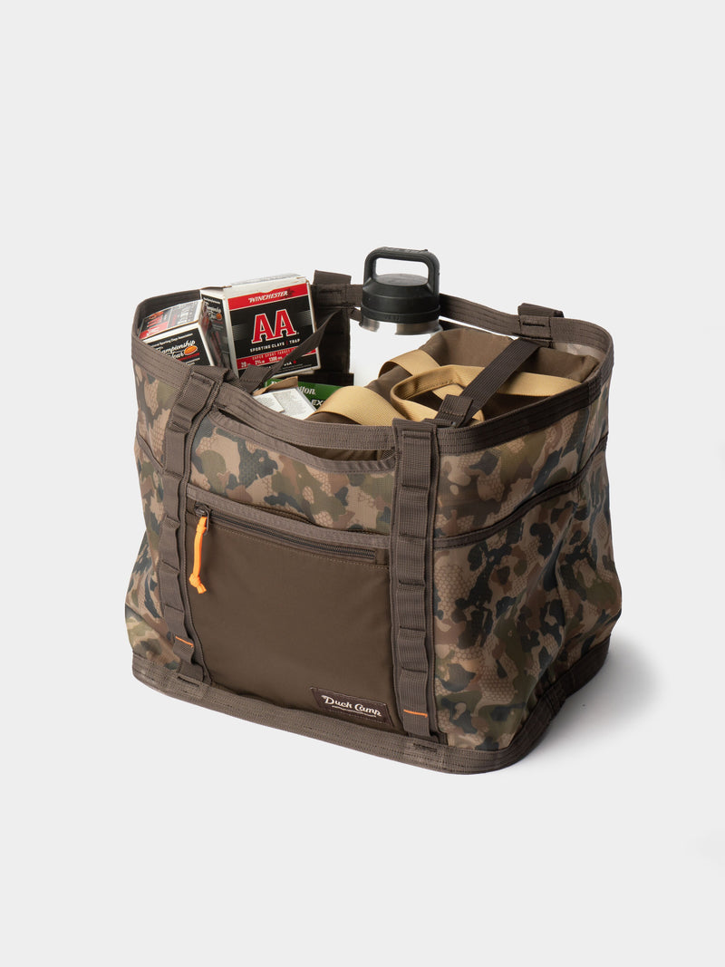 The Duck Camp Mesh Gear Tote features a camouflage pattern and holds ammunition boxes, a water bottle, and tan straps, all displayed against a white background.
