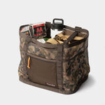 The Duck Camp Mesh Gear Tote is a camo-patterned, water-resistant bag with tan straps, shown on a white background and filled with boxes of AA shotgun shells and a water bottle.