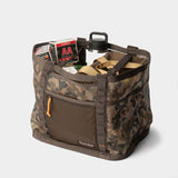 The Duck Camp Mesh Gear Tote features a camouflage pattern and holds ammunition boxes, a water bottle, and tan straps, all displayed against a white background.