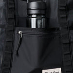 A black water bottle sits in the mesh pocket of the Duck Camp Mesh Gear Tote, a black outdoor storage bag featuring a zippered front pocket and branded label.