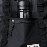 A black water bottle sits in the mesh pocket behind the zippered front pocket of a black Duck Camp Mesh Gear Tote.