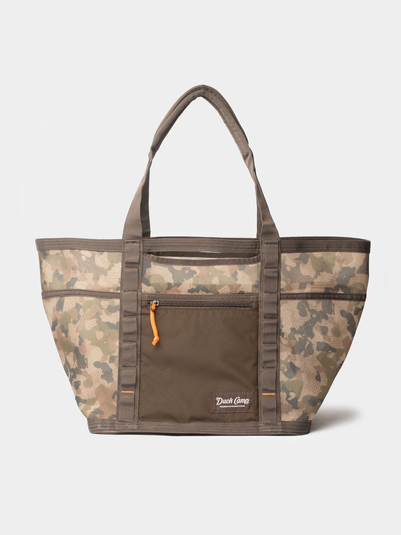 The Duck Camp Mesh Gear Tote is a camouflage-patterned, water-resistant tote with two handles, a front zippered pocket with orange pull, and a "Duck Camp" logo—ideal for carrying outdoor gear on your next adventure.