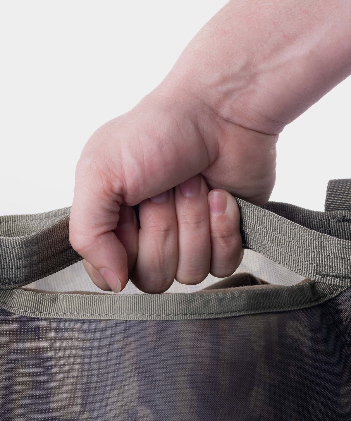 A close-up of a hand gripping the handle of Duck Camp's Mesh Gear Tote, a green and brown camouflage heavy-duty bag, shown against a plain light background.