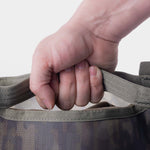 A close-up of a hand gripping the handle of the Duck Camp Mesh Gear Tote, a heavy-duty bag featuring a camouflage pattern, set against a plain white background.