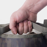A close-up of a hand gripping the handle of Duck Camp's Mesh Gear Tote, a green and brown camouflage heavy-duty bag, shown against a plain light background.