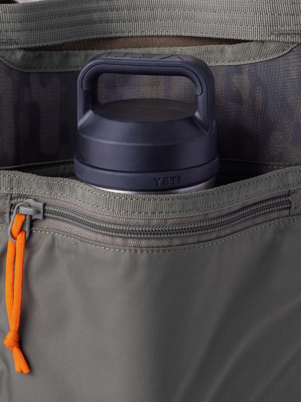 A close-up of a gray Yeti bottle partially tucked inside a Duck Camp Mesh Gear Tote's zippered pocket with an orange zipper pull.