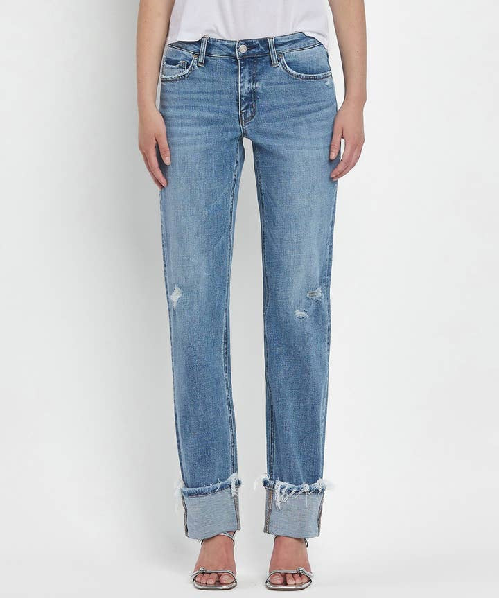 A person wears Vervet's Mid Rise Cuffed Straight Jean in light blue with distressed hems and minor rips, styled with a white top and open-toe heels against a plain background.