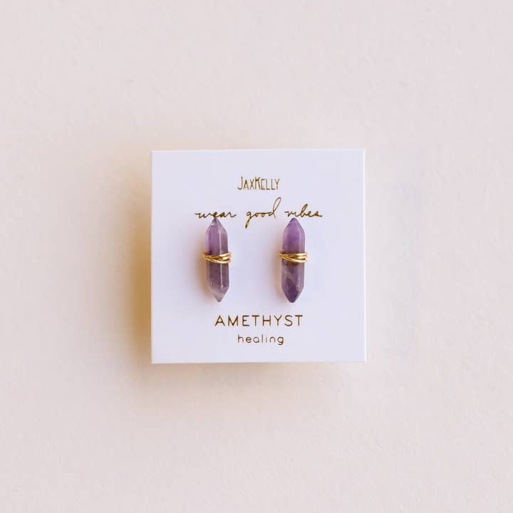A pair of Mineral Point Amethyst Gold earrings by JaxKelly, featuring double point amethyst crystals wrapped in gold wire, displayed on a white card labeled "JaxKelly" and "Amethyst healing.
