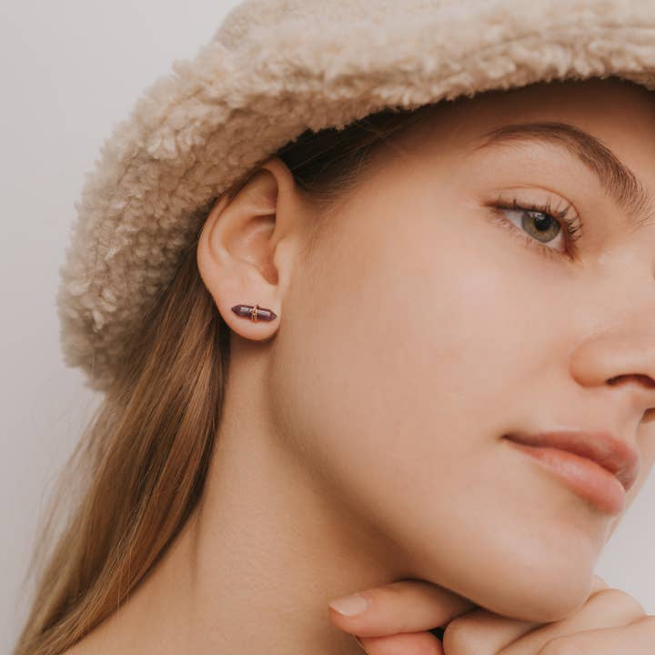 A woman in a beige fuzzy hat wears Mineral Point Amethyst Gold earrings by JaxKelly, resting her chin on her hand and shown in profile against a plain background.