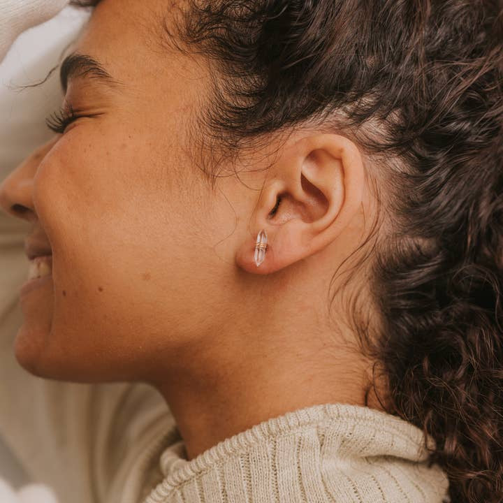 Close-up of a woman with curly hair smiling, wearing JaxKelly Mineral Point Clear Quartz earrings and a beige knit sweater.