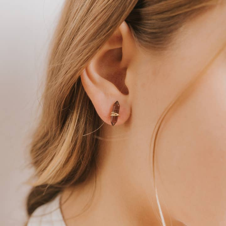 Close-up of a woman's ear with light brown hair, wearing the Mineral Point Smoky Quartz earring by JaxKelly—leaf-shaped, brown, hypoallergenic, and featuring a gold horizontal accent.