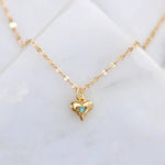 The Mesa Blue Mini Opal Heart Necklace features an 18K gold plated chain and a small gold heart pendant with a turquoise stone center, displayed on a white marble surface.
