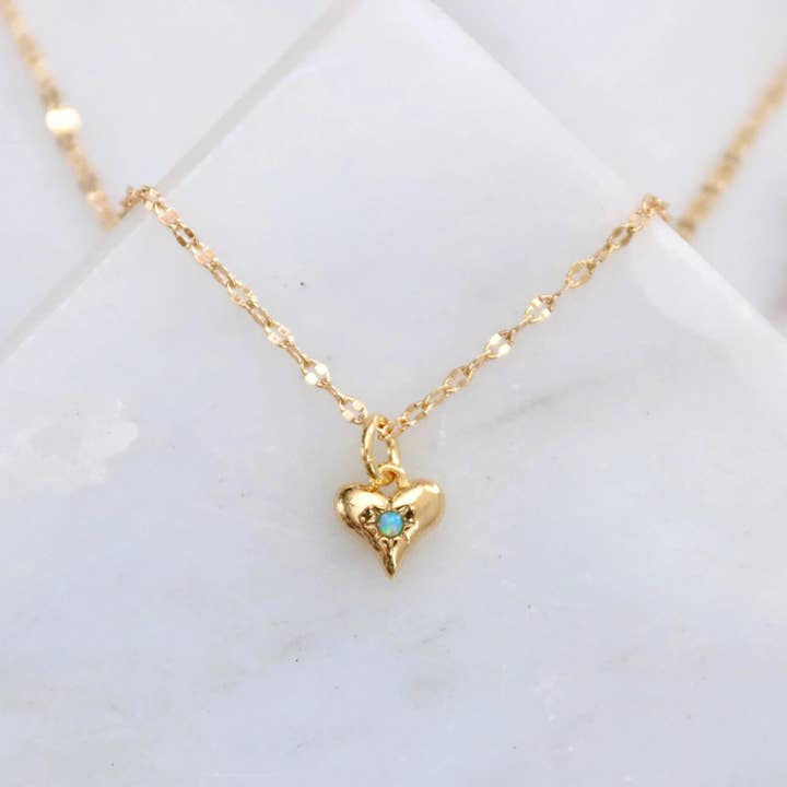 The Mesa Blue Mini Opal Heart Necklace is an 18K gold plated chain with a small heart-shaped pendant featuring a central opal, shown on a white marble background.