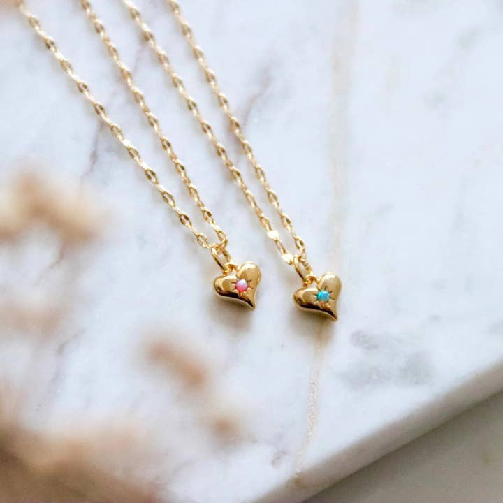 Two Mesa Blue Mini Opal Heart Necklaces—one pink opal, one blue—feature 18K gold plating and heart-shaped pendants, each with a small gem, elegantly displayed on a white marble surface.