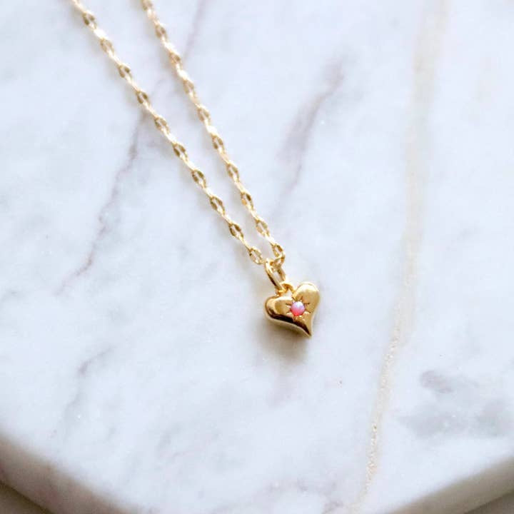 The Mesa Blue Mini Opal Heart Necklace features an 18K gold plated chain and a small pink stone heart pendant, reminiscent of a delicate pink opal, shown on a white marble surface.
