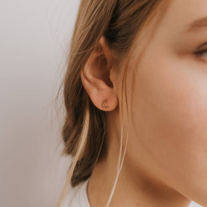 Close-up of a woman's ear wearing JaxKelly's Minimalist Moon Gold Earrings. Her hair is tucked behind her ear, and the background is plain.