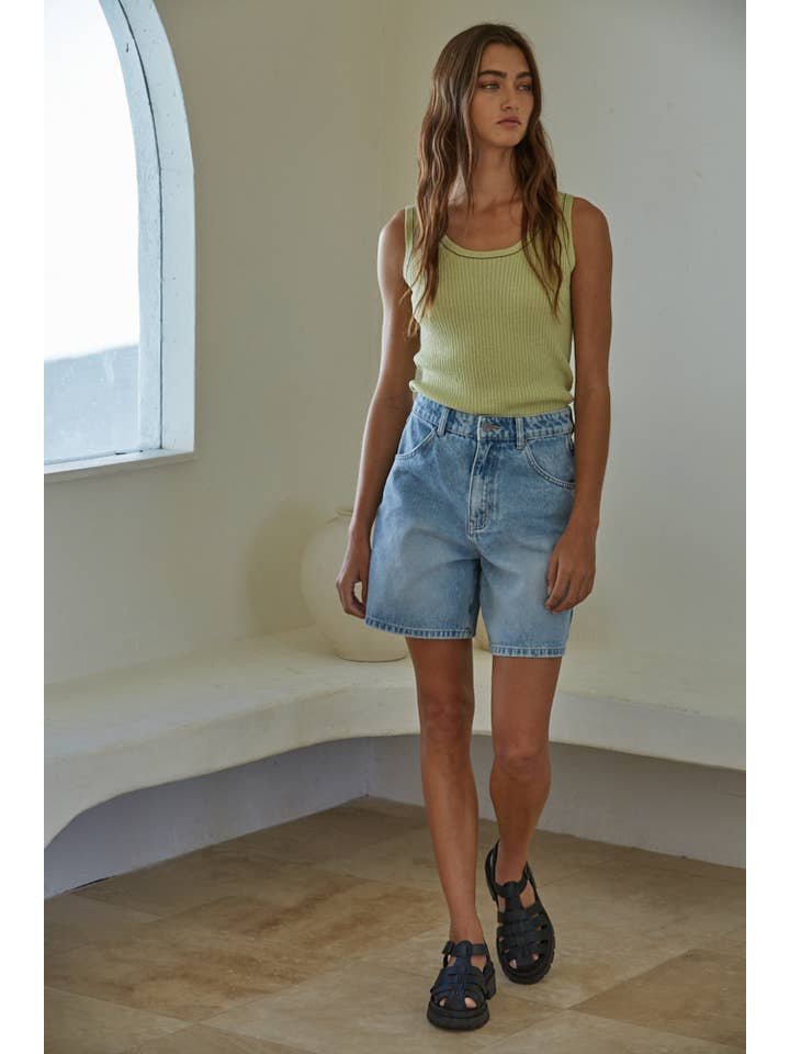 A woman stands indoors in a light yellow Mira Sleeveless Top by By Together, denim shorts, and black sandals, with a window in the background.