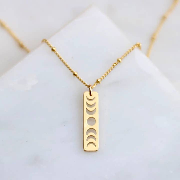 The Moon Phase Necklace by Mesa Blue features a rectangular 18K gold-plated pendant with cut-out moon phases, displayed on a white marble surface.