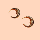 The Moonstruck Hoops by Peter and June are 8k gold plated crescent moon-shaped earrings with a face design, made from ethically sourced materials and shown on a peach background.