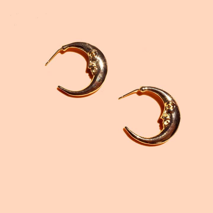 The Moonstruck Hoops by Peter and June are 8k gold plated crescent moon-shaped earrings with a face design, made from ethically sourced materials and shown on a peach background.