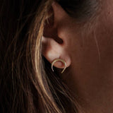 Close-up of a person wearing the Naja Brass Studs by CommonForm, a small crescent-shaped earring, with brown hair partially visible.