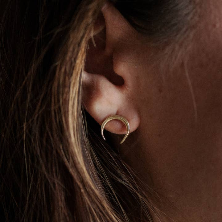 Close-up of a person wearing the Naja Brass Studs by CommonForm, a small crescent-shaped earring, with brown hair partially visible.