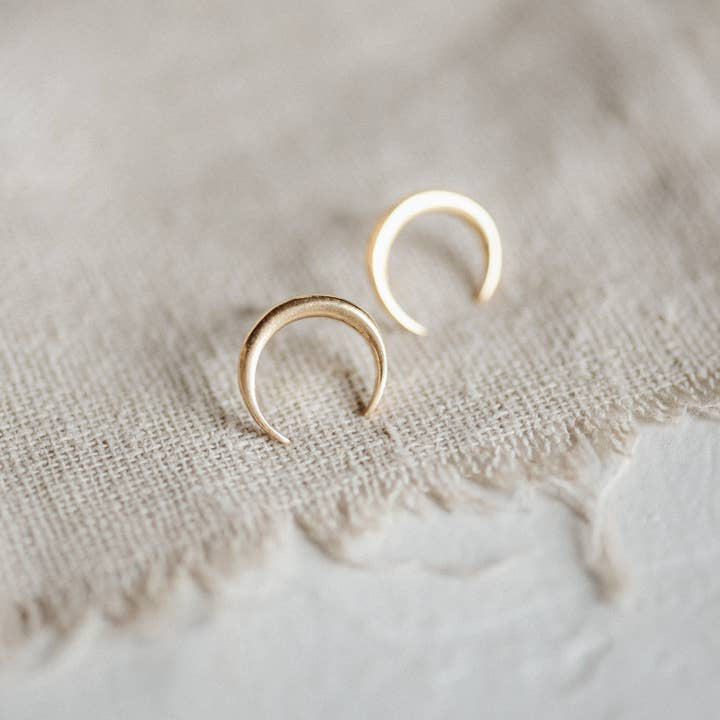 Naja Brass Studs by CommonForm—a pair of small crescent earrings with a 24K gold plate—rest on a textured beige fabric surface.