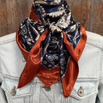 A Navy and Rust Floral Paisley Wild Rag by Rodeo Drive is tied around the neck of a person in a light denim jacket, set against a wooden background.