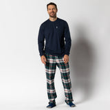 A man stands smiling in the Navy Lounge Set by Duck Head—featuring an ultra-soft cotton tee, plaid green-red-white flannel pants, and blue socks. The perfect gift look against a plain white background.