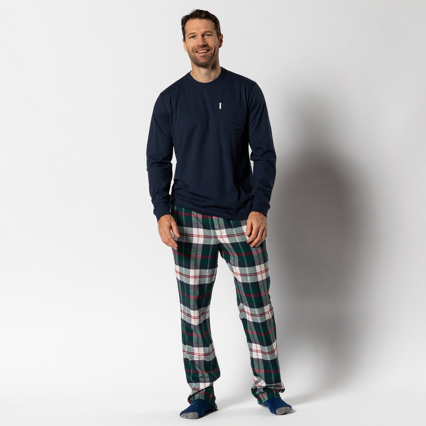 A man stands smiling in the Navy Lounge Set by Duck Head—featuring an ultra-soft cotton tee, plaid green-red-white flannel pants, and blue socks. The perfect gift look against a plain white background.
