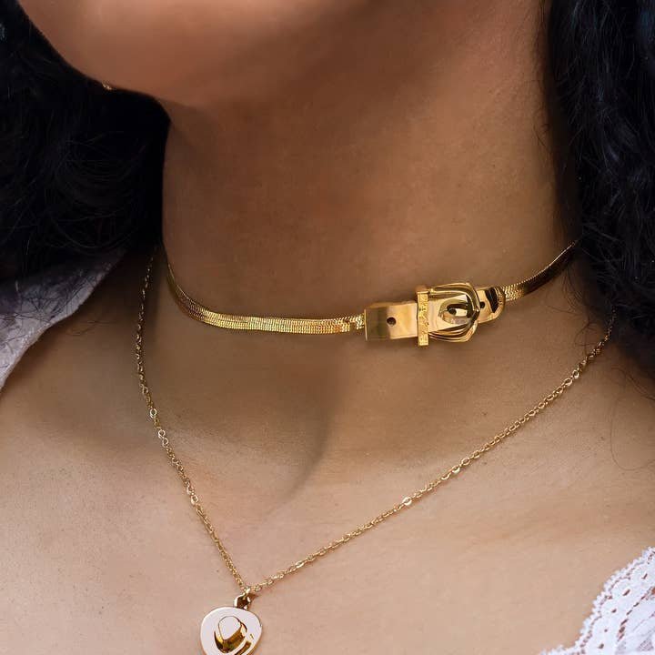 A person wears the Peter and June Brooks Buckle Choker, an 18k gold plated necklace with a buckle design, paired with an adjustable gold pendant necklace featuring a white accent.
