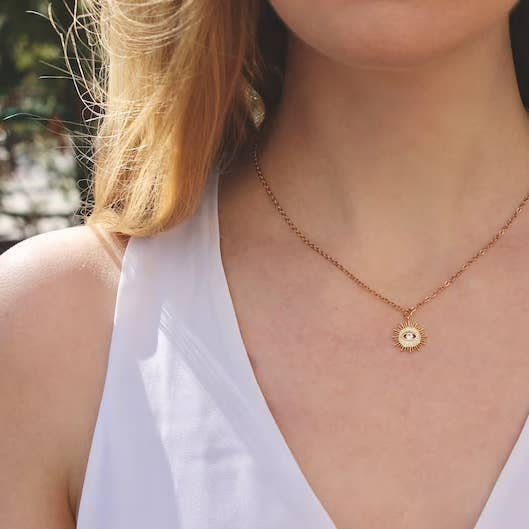 A person wearing a white sleeveless top and the Vintage Acorn Necklace | Crystal Eye Sunburst featuring a 16K gold plated sun-shaped pendant with a gold filled spring clasp.