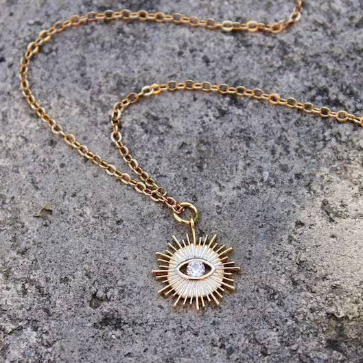 The Vintage Acorn Necklace | Crystal Eye Sunburst features a 16K gold plated sunburst pendant with a central eye and clear gemstone. Nickel and lead free for comfort, it's shown on a rough gray surface.