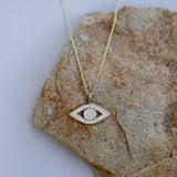 JaxKelly Cz Evil Eye Necklace: gold plated, tarnish free for lasting shine, featuring an eye-shaped pendant with clear stones. Displayed on textured brown stone.