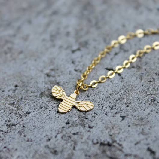 The Necklace | Gold Bee by Vintage Acorn, featuring a 16K gold plated pendant, is displayed on a rough gray concrete surface.