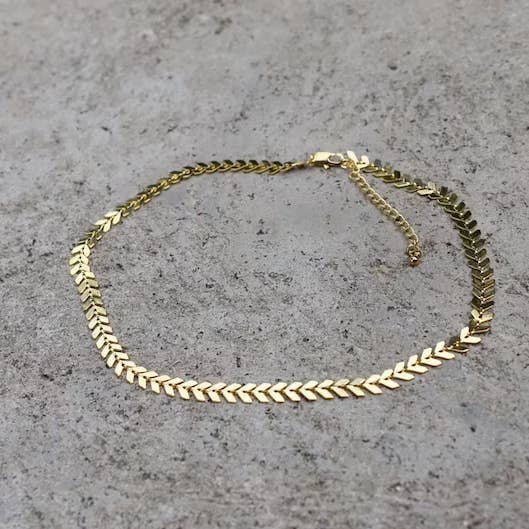 The Gold Chevron Necklace by Vintage Acorn is displayed flat on a gray textured surface, featuring a 16k gold-plated herringbone pattern.