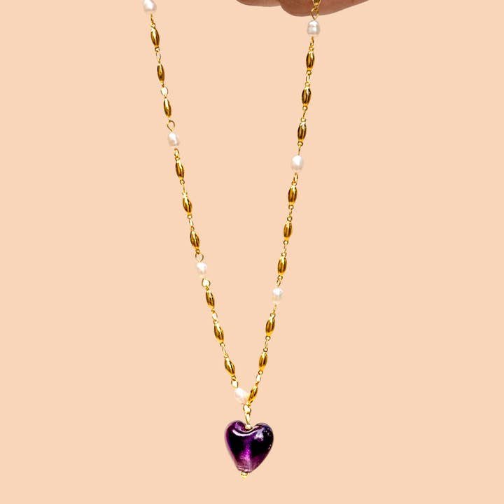A hand holds the Peter and June Jill Necklace, a 24k gold plated piece with freshwater pearls and a purple heart-shaped pendant, against a light peach background.