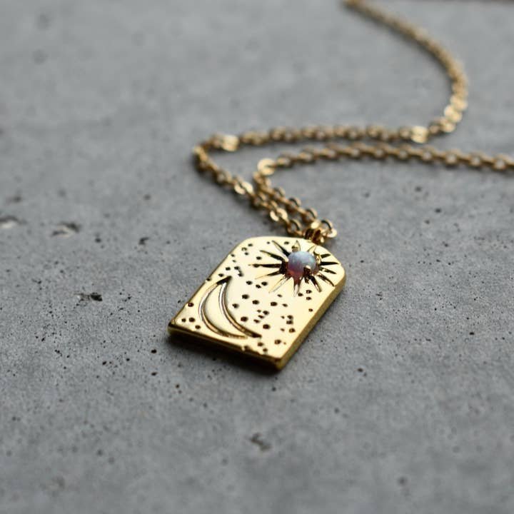 The Opal Starry Night Necklace by Vintage Acorn features a 16K gold plated rectangular pendant with an engraved crescent moon, starburst design, opal stone on a textured surface, and a gold filled spring clasp. Nickel and lead free.