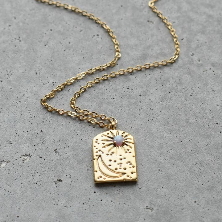 The Necklace | Opal Starry Night by Vintage Acorn features a 16K gold plated rectangular pendant with crescent moon, starburst, textured dots, and opal gemstone. Nickel and lead free. Shown on a gray concrete surface.