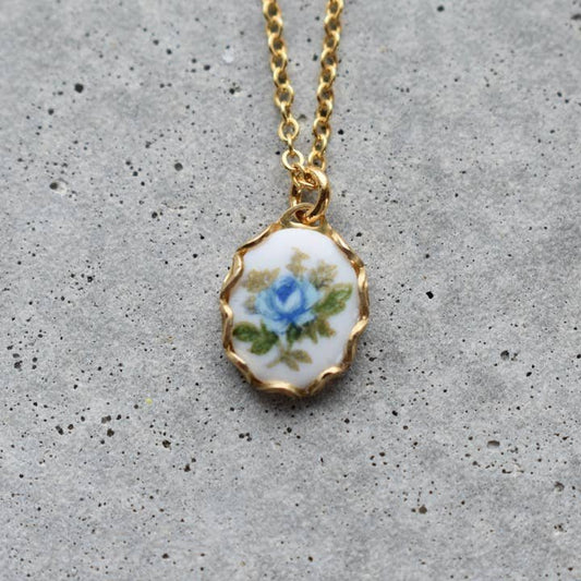 Vintage Acorn's Necklace | Vintage Floral features a 16K gold plated chain and pendant with a hand-painted blue rose and green leaves on white. Nickel and lead free, with a gold filled spring clasp. Displayed on gray concrete.