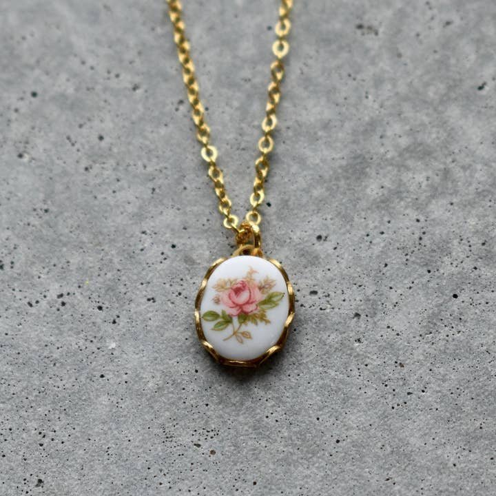 Vintage Acorn's "Necklace | Vintage Floral | Vintage Acorn" features a 16K gold plated pendant with a pink rose and green leaves on white. Nickel and lead free for comfort, shown on a gray textured surface.