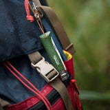 An Opinel No. 08 Colorama Forest Green folding knife is secured by a leather loop to a blue and red backpack, whose surface is dotted with water droplets.