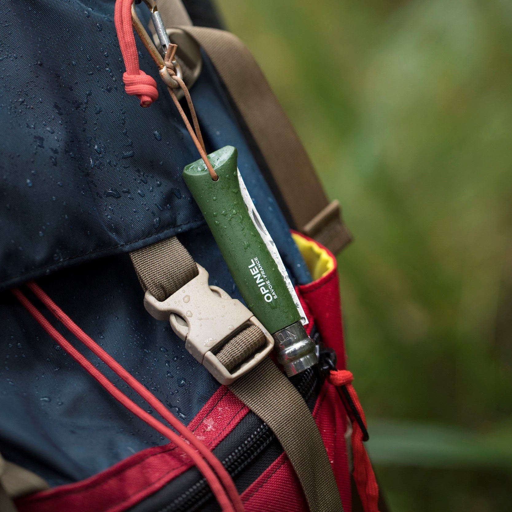 An Opinel No. 08 Colorama Forest Green folding knife is secured by a leather loop to a blue and red backpack, whose surface is dotted with water droplets.