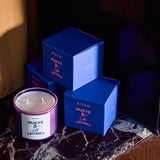 A purple Roen Nueve Leones coconut soy wax candle with a white label rests on marble beside three blue boxes of Roen hand-poured candles.
