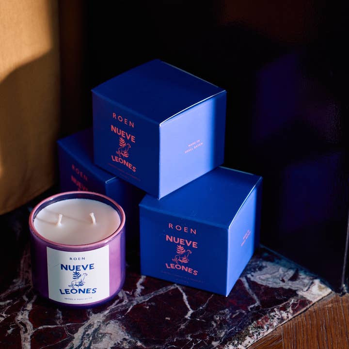 A Nueve Leones candle by Roen, crafted from coconut soy wax in purple, sits on a marble surface next to three stacked blue boxes.